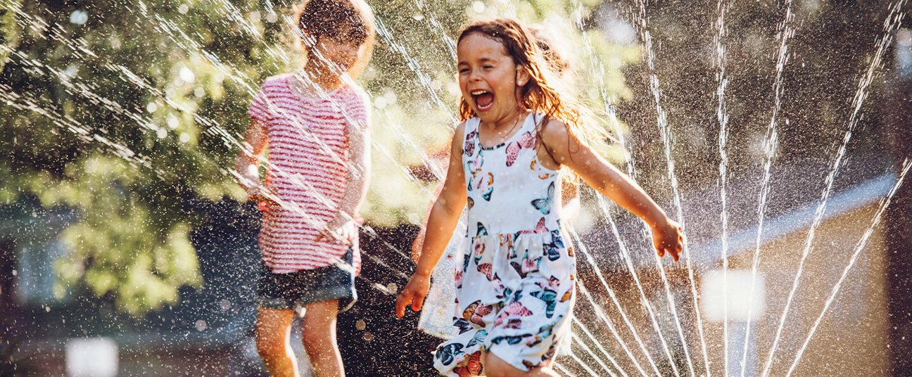 Zwei lachende Kinder spielen an einem heißen Sommertag im Wasserstrahl eines Rasensprengers