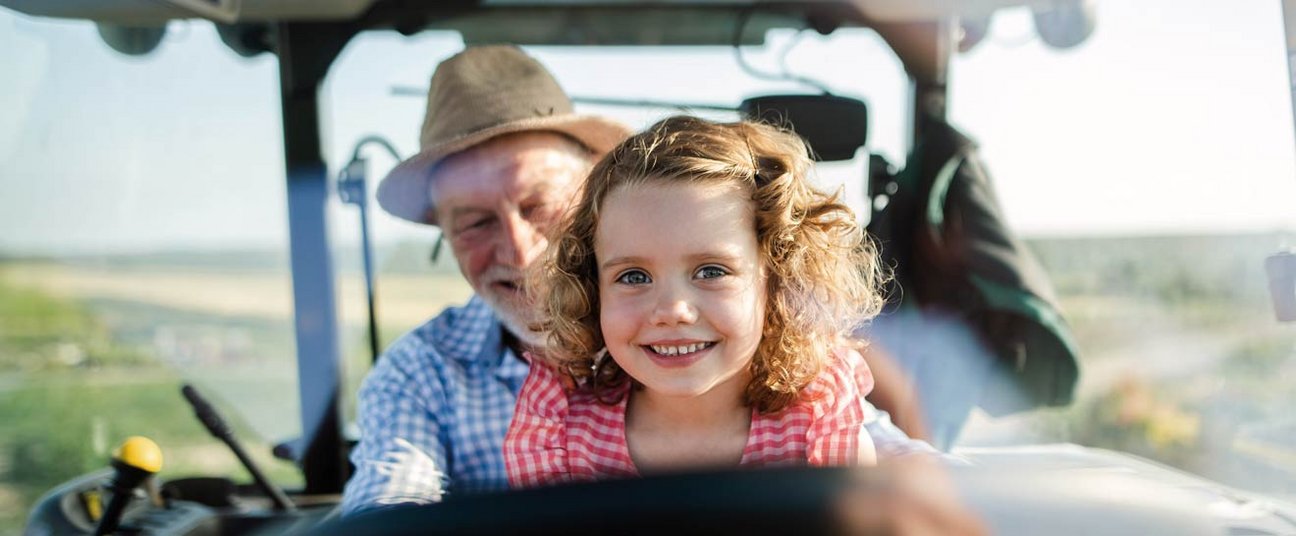 Das Bild zeigt ein junges Mädchen, wie es fröhlich mit dem Opa Traktor fährt.