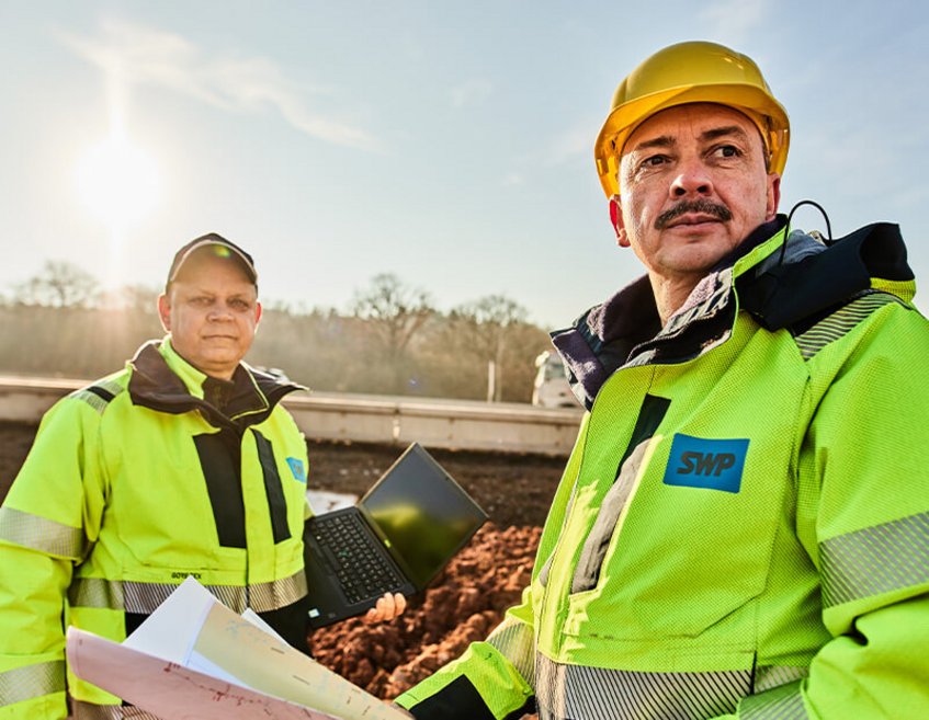 Zwei SWP-Fachkräfte auf Baustelle mit Laptop und Plänen, im Hintergrund eine Straße und Sonnenlicht