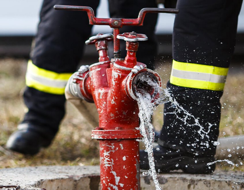 Wasser sprudelt aus rotem Hydranten, Feuerwehrleute in Einsatzkleidung im Hintergrund