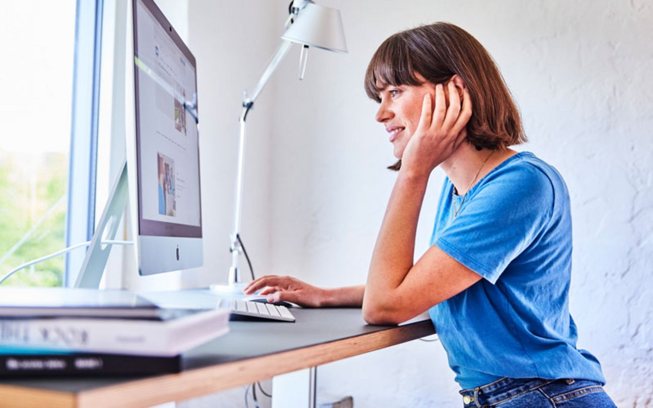 Junge Frau steht am Schreibtisch und arbeitet an einem Desktop-PC