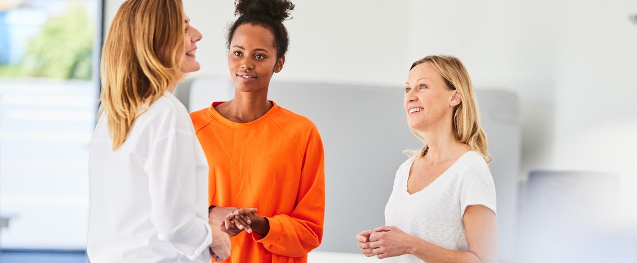 Drei Frauen stehen in hellem Raum zusammen und unterhalten sich.