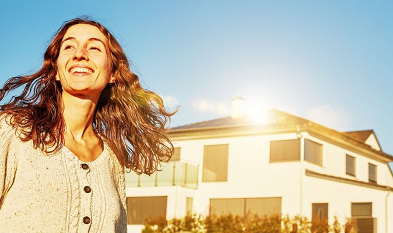 Das Bild zeigt eine junge Frau die der Sonne entgegen lacht, Hinter Ihr ist ein Haus mit einer PV-Anlage auf dem Dach