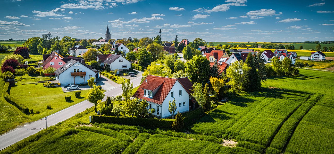 Luftaufnahme eines idyllischen Dorfs mit Einfamilienhäusern, Feldern und Kirche bei sonnigem Wetter