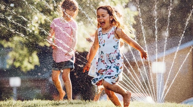 Zwei lachende Kinder spielen an einem heißen Sommertag im Wasserstrahl eines Rasensprengers