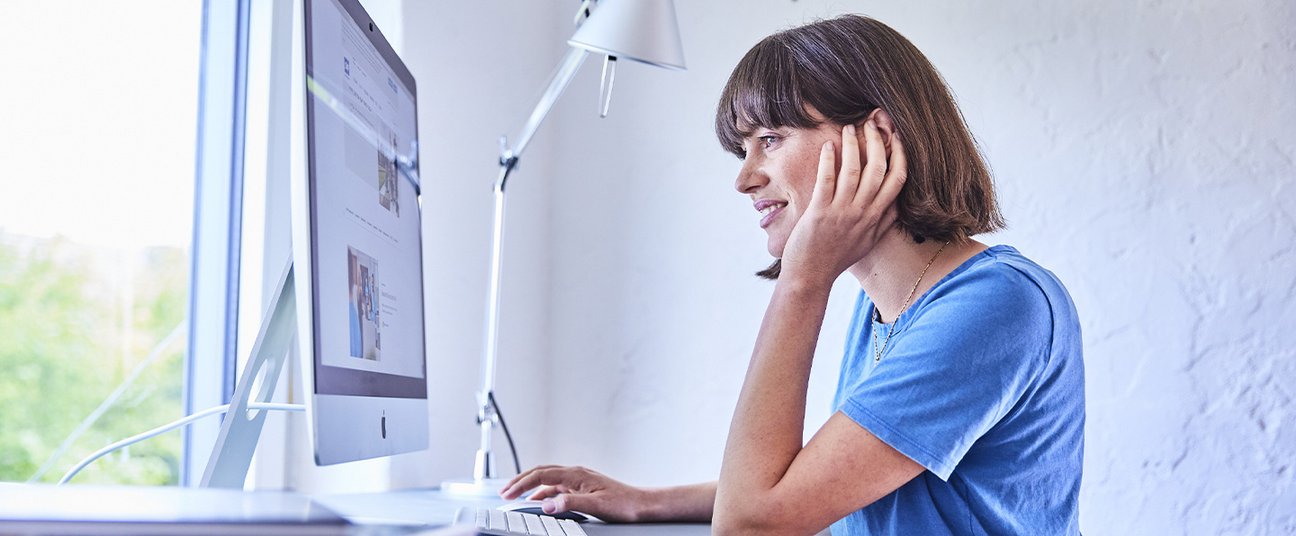 Junge Frau sitzt an einem Schreibtisch und arbeitet an einem Desktop-Computer in modernem, hellem Raum.