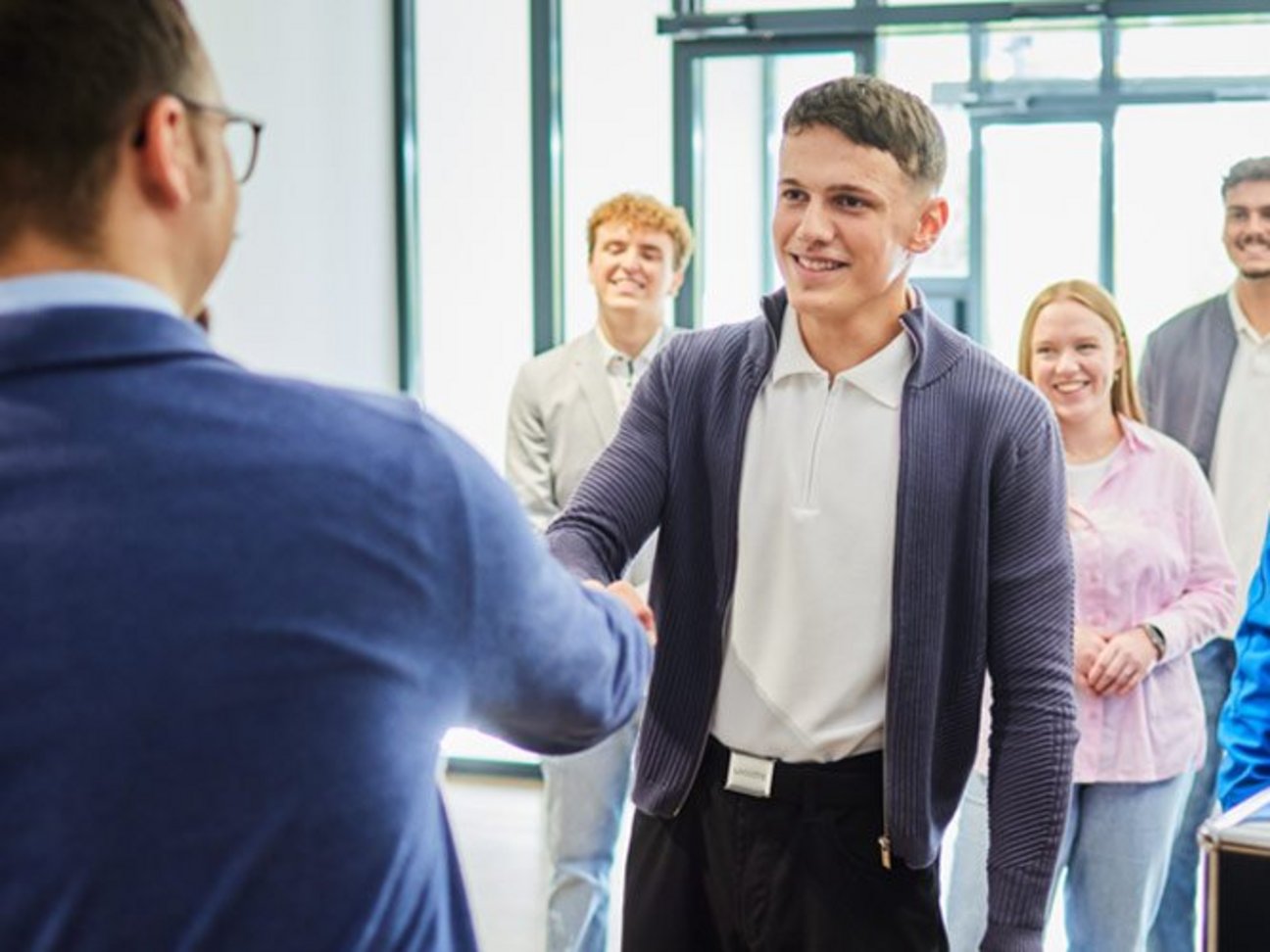 Das Bild zeigt einen jungen Mann, der von eine anderen Mann begrüßt wird und weitere fröhliche Menschen stehen hinter diesem jungen Mann. 