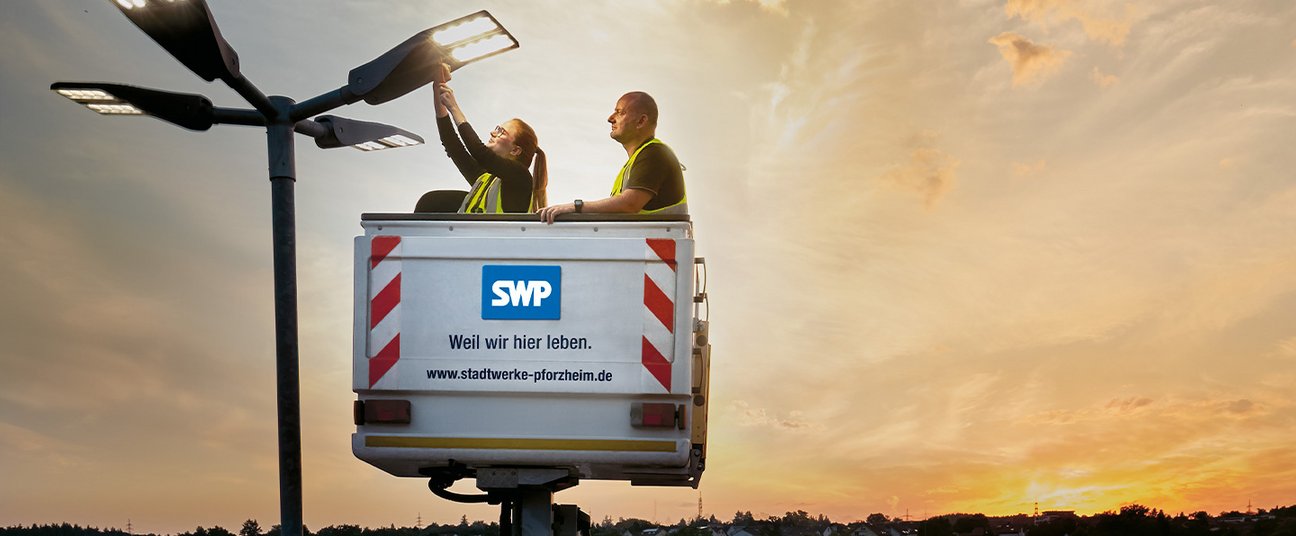Das Einstiegsbild zeigt 2 Kollegen, die auf einem Steiger eine Straßenlaterne reparieren
